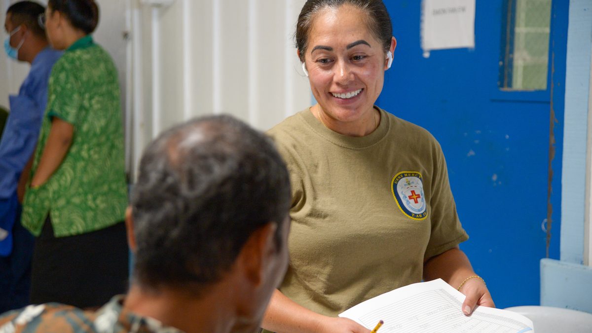 A smiling health worker holds a printed registration form and a pencil while she talks to a seated patient, seen from behind, in a busy clinic waiting area.