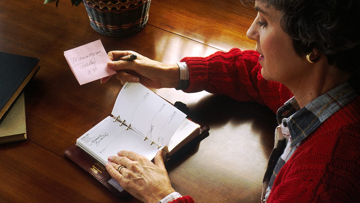 A woman at a wooden desk holds a handwritten appointment note over an open paper diary